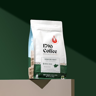 A bag of 1790 Coffee Specialty Coffee displayed on a green background. The coffee bag is white in the upper part with green at the bottom. It has labels indicating 'Signature Roast,' 'Single Origin Mexico,' 'Whole Bean,' and 'Medium Roast.' The design includes graphical elements of a building and a flame logo, along with text describing the flavors as 'Graham Cracker, Milk Chocolate, Orange.' The weight of the coffee is 12 oz (340g) and it is labeled as 100% Organic Arabica Coffee.