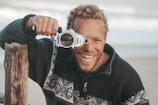 A person with curly hair and a beard is smiling while holding a camera with a unique housing to their eye. They are wearing a sweater with a geometric pattern and there's a wooden post nearby. The background is a soft-focus beach setting, suggesting a relaxed environment.