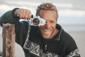 A person with curly hair and a beard is smiling while holding a camera with a unique housing to their eye. They are wearing a sweater with a geometric pattern and there's a wooden post nearby. The background is a soft-focus beach setting, suggesting a relaxed environment.