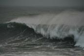 A large ocean wave surges with force, topped by a misty spray that creates a dramatic scene. The sea appears turbulent, with shades of green and grey indicating a stormy atmosphere.