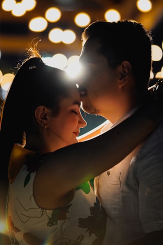 A couple shares an intimate moment with one person gently kissing the other's forehead. The setting is dimly lit with a bokeh effect in the background, suggesting lights or a cityscape at night. The scene feels romantic and serene, with soft lighting highlighting their faces.