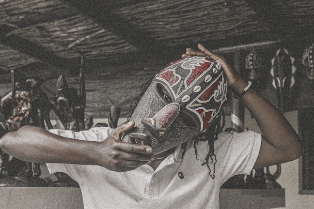 A person wearing a decorative tribal mask, embellished with red, black, and white patterns, holds it firmly in place. The background includes several wooden carved statues, giving a rustic and culturally rich atmosphere.