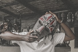 A person wearing a decorative tribal mask, embellished with red, black, and white patterns, holds it firmly in place. The background includes several wooden carved statues, giving a rustic and culturally rich atmosphere.