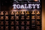 A wall with multiple electrical panels is displayed. Above the panels, a neon sign reads 'TOALETY', which suggests a public restroom indicator. The setup is industrial with a utilitarian aesthetic, featuring dark tones and exposed wiring.