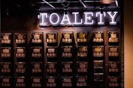 A wall with multiple electrical panels is displayed. Above the panels, a neon sign reads 'TOALETY', which suggests a public restroom indicator. The setup is industrial with a utilitarian aesthetic, featuring dark tones and exposed wiring.