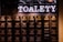 A wall with multiple electrical panels is displayed. Above the panels, a neon sign reads 'TOALETY', which suggests a public restroom indicator. The setup is industrial with a utilitarian aesthetic, featuring dark tones and exposed wiring.