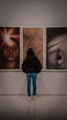 A person is standing in front of three abstract portraits displayed on a wall. The person is viewing the artworks closely, suggesting interest and engagement. The portraits each have distinct features, with the left one showing a face illuminated by a bright light, the center one having a blurred, warm-toned appearance, and the right one depicting a prominent eye.