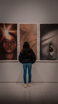 A person is standing in front of three abstract portraits displayed on a wall. The person is viewing the artworks closely, suggesting interest and engagement. The portraits each have distinct features, with the left one showing a face illuminated by a bright light, the center one having a blurred, warm-toned appearance, and the right one depicting a prominent eye.
