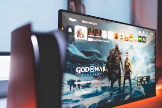 A widescreen TV or monitor displays the main screen of a video game titled 'God of War Ragnarök'. The background features a wintery, mystical landscape with characters wearing armor. The room is dimly lit, adding a dramatic effect.