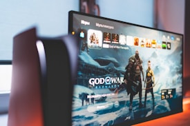 A widescreen TV or monitor displays the main screen of a video game titled 'God of War Ragnarök'. The background features a wintery, mystical landscape with characters wearing armor. The room is dimly lit, adding a dramatic effect.