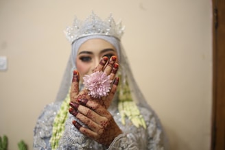 A bride is holding a pink flower in her hands, which are decorated with intricate henna designs and dark nail polish. She is dressed in an elaborate, shimmering outfit with a matching ornate crown. The style of the attire suggests a traditional or cultural dress. The background is simple and undistracting, focusing attention on the bride.