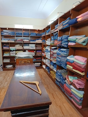 The image depicts the interior of a store with numerous shelves lining the walls, all filled with neatly folded fabrics of various colors and patterns. A long wooden table stands in the middle of the room, with a wooden hanger placed on it. The setting appears organized and well-lit, with several light fixtures illuminating the space.