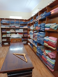 The image depicts the interior of a store with numerous shelves lining the walls, all filled with neatly folded fabrics of various colors and patterns. A long wooden table stands in the middle of the room, with a wooden hanger placed on it. The setting appears organized and well-lit, with several light fixtures illuminating the space.