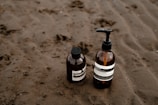 Two brown glass bottles with pump dispensers are placed on a sandy beach. The bottles have white labels with black text and appear to contain liquid products, likely skincare or toiletries.