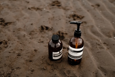 Two brown glass bottles with pump dispensers are placed on a sandy beach. The bottles have white labels with black text and appear to contain liquid products, likely skincare or toiletries.
