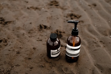 Two brown glass bottles with pump dispensers are placed on a sandy beach. The bottles have white labels with black text and appear to contain liquid products, likely skincare or toiletries.