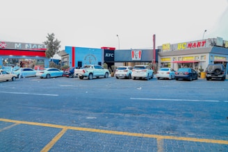 A parking lot in front of several stores including a KFC, M Mart, and BC Mart with parked cars and a clear sky overhead. The buildings have vibrant colors and the scene depicts a typical urban shopping area.