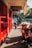 A narrow street features bright red doors and a signboard with the word 'LARIS.' People are seen walking and engaging in activities. Motorcycles are parked along the street, while bags and other items are stacked near the curb. The scene is lit by sunlight, casting shadows on the ground.