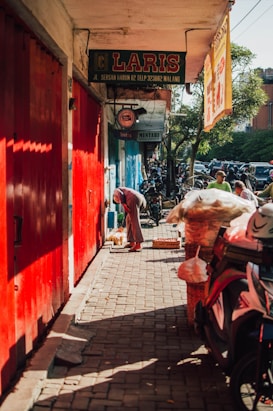 A narrow street features bright red doors and a signboard with the word 'LARIS.' People are seen walking and engaging in activities. Motorcycles are parked along the street, while bags and other items are stacked near the curb. The scene is lit by sunlight, casting shadows on the ground.