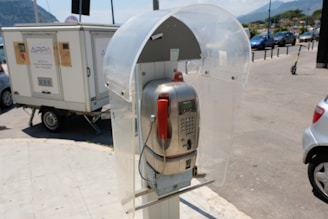 A public payphone with a silver metallic body and a red handset is housed within a transparent, protective booth. The phone is situated outdoors, with a street scene in the background showing parked cars and a small trailer. The environment appears sunny, casting clear shadows on the ground.