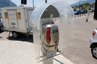 A public payphone with a silver metallic body and a red handset is housed within a transparent, protective booth. The phone is situated outdoors, with a street scene in the background showing parked cars and a small trailer. The environment appears sunny, casting clear shadows on the ground.