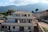A large, two-story residential building with a slightly worn appearance, surrounded by greenery and mountains in the background. The sky is partly cloudy with sunlight casting gentle shadows on the structure.