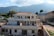A large, two-story residential building with a slightly worn appearance, surrounded by greenery and mountains in the background. The sky is partly cloudy with sunlight casting gentle shadows on the structure.