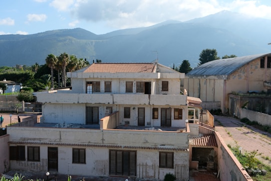 A large, two-story residential building with a slightly worn appearance, surrounded by greenery and mountains in the background. The sky is partly cloudy with sunlight casting gentle shadows on the structure.