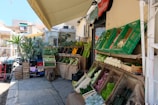 An outdoor market stall is filled with a variety of fresh vegetables, including lettuce, zucchinis, eggplants, and some herbs. The produce is displayed in wooden crates and baskets with some burlap sacks. Above, an awning provides shade and there are other market stalls and buildings in the background.