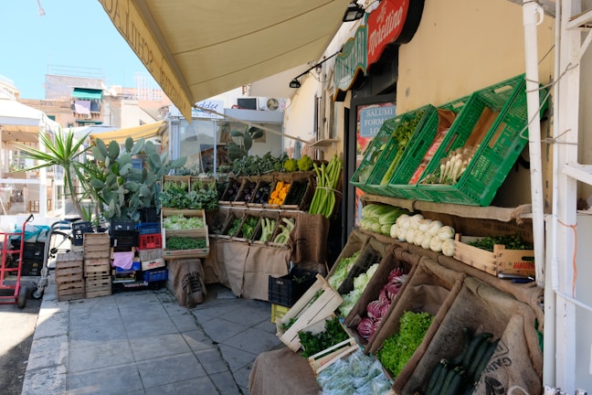 An outdoor market stall is filled with a variety of fresh vegetables, including lettuce, zucchinis, eggplants, and some herbs. The produce is displayed in wooden crates and baskets with some burlap sacks. Above, an awning provides shade and there are other market stalls and buildings in the background.