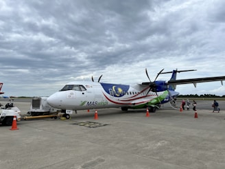 An airplane on the tarmac with MASwings livery, featuring the Malaysian flag colors and logo. The aircraft has propellers and is parked near a ground vehicle. A line of passengers is visible boarding the plane using a small set of mobile stairs. Orange traffic cones mark the area around the aircraft on the cloudy day.