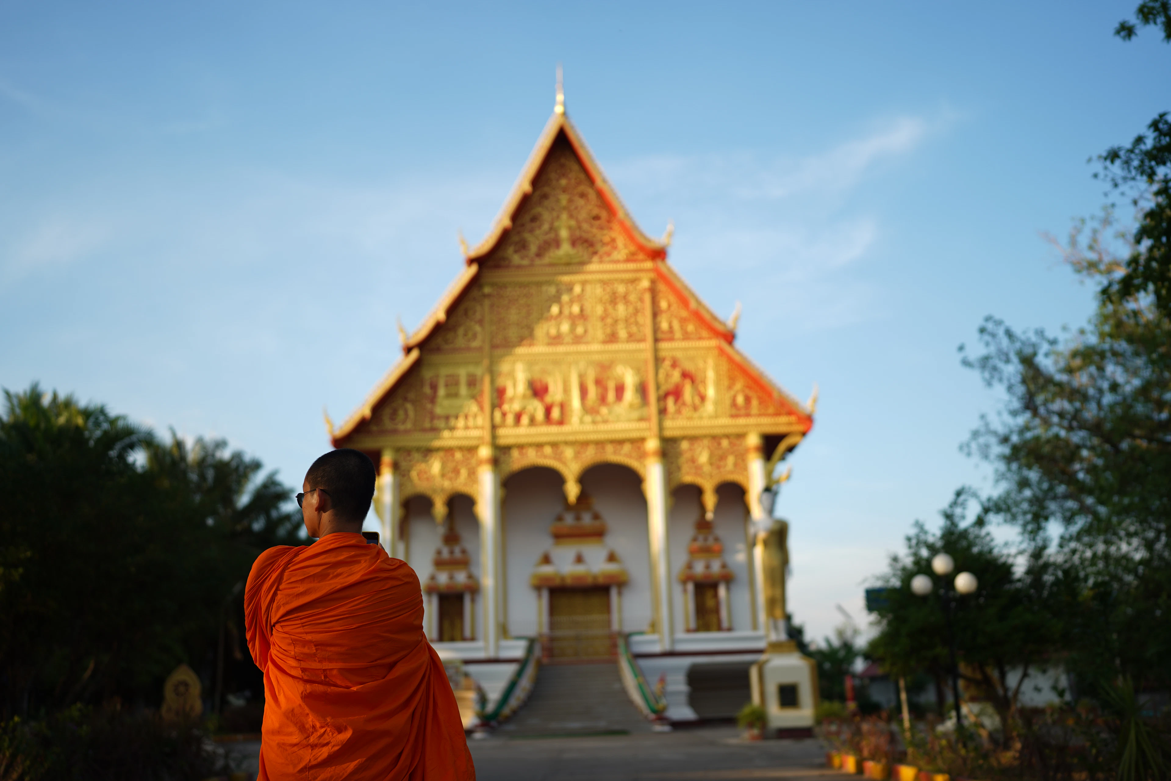A monk in saffron robes standing before the gilded gable and tiered roof of a Lao temple hall in Vientiane, the low evening sun setting the gold leaf alight against a clear blue sky