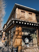 A rustic wooden building with a shop at street level displaying various colorful bags and decorative items. The building features wooden textures and small windows on the upper floor, with a slanted roof. There is a small black signboard on the wall and a pedestrian pathway alongside lined with trees.