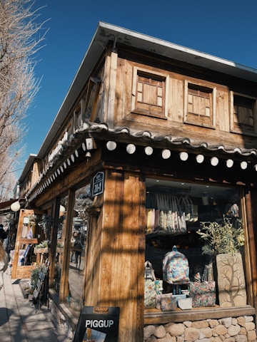 A rustic wooden building with a shop at street level displaying various colorful bags and decorative items. The building features wooden textures and small windows on the upper floor, with a slanted roof. There is a small black signboard on the wall and a pedestrian pathway alongside lined with trees.