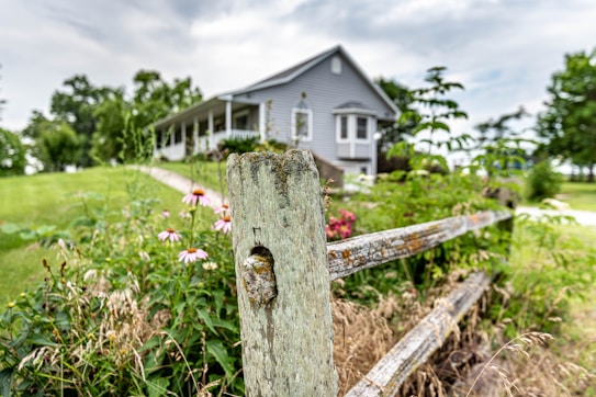 A rustic wooden fence in the foreground with weathered texture stands in front of a lush garden filled with greenery and pink flowers. In the background, a quaint grey house with a porch is partially visible, surrounded by green lawns and trees under a cloudy sky.