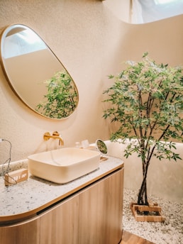 A modern bathroom with a natural aesthetic features a large oval mirror mounted on a textured wall. A rectangular vessel sink is placed on a speckled countertop, and gold-colored faucet fixtures add elegance. An indoor plant is set in a wooden planter, surrounded by small white pebbles that cover part of the floor.