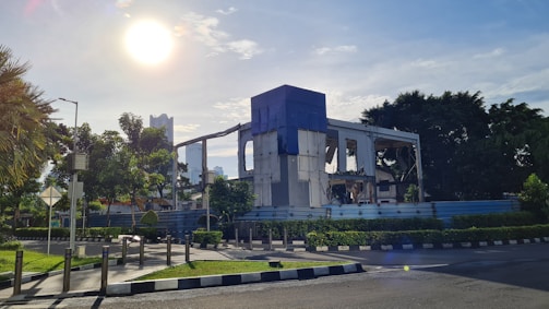 A partially demolished building stands surrounded by greenery and a clear sky. The sun is bright in the background, casting light over the scene. Trees and well-maintained bushes line the street, and the road is bordered by a low fence.