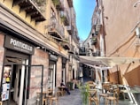 A narrow street with a small Italian restaurant called Ponticello located on the left. Several balconies with plants, lamps, and decor are visible on the buildings. There are chairs and tables outside, with one person sitting near the restaurant. The street is lined with buildings that have colorful walls and various window designs. There is a canopy covering some of the outdoor seating area, and the sky is clear and blue.