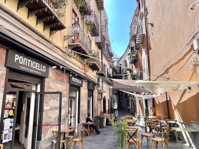 A narrow street with a small Italian restaurant called Ponticello located on the left. Several balconies with plants, lamps, and decor are visible on the buildings. There are chairs and tables outside, with one person sitting near the restaurant. The street is lined with buildings that have colorful walls and various window designs. There is a canopy covering some of the outdoor seating area, and the sky is clear and blue.