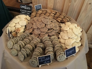 A large assortment of cookies is displayed on a table, organized by type. There are clusters of peanut butter, mint chocolate chip, and sugar cookies, each with a small sign indicating the flavor. The cookies are arranged in concentric circles on a beige tablecloth, set against a wooden backdrop.