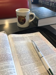An open book with handwritten notes and a pen rests on a table. A mug with a 'Waffle House Coffee' logo and a closed book are nearby. The setting appears to be a casual dining area with a padded red seat in the background.