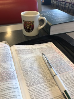An open book with handwritten notes and a pen rests on a table. A mug with a 'Waffle House Coffee' logo and a closed book are nearby. The setting appears to be a casual dining area with a padded red seat in the background.