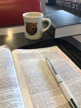 An open book with handwritten notes and a pen rests on a table. A mug with a 'Waffle House Coffee' logo and a closed book are nearby. The setting appears to be a casual dining area with a padded red seat in the background.