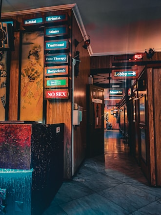 A hallway with wooden walls and various illuminated signs in neon colors pointing to different services such as 'Shiatsu', 'Traditional', and 'Face Therapy'. There is a colorful painting of a Geisha on one wall. The floor is marble, reflecting the lights, and the atmosphere is somewhat dim.
