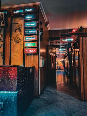 A hallway with wooden walls and various illuminated signs in neon colors pointing to different services such as 'Shiatsu', 'Traditional', and 'Face Therapy'. There is a colorful painting of a Geisha on one wall. The floor is marble, reflecting the lights, and the atmosphere is somewhat dim.