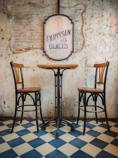 A rustic café setting with a small round table and two wooden chairs on a blue and white checkered floor. The wall has a textured, vintage look with a large sign reading 'Artisan des Glaces' and advertises ice cream and sorbet.
