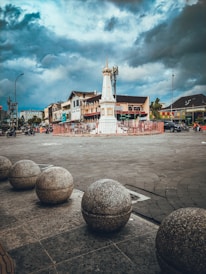 A historical monument stands in the center of a roundabout, surrounded by stone spheres that line the foreground. The sky is overcast with dramatic clouds. In the background, buildings and a few vehicles, including motorcycles, are visible. The atmosphere is a mix of urban life and historical architecture.