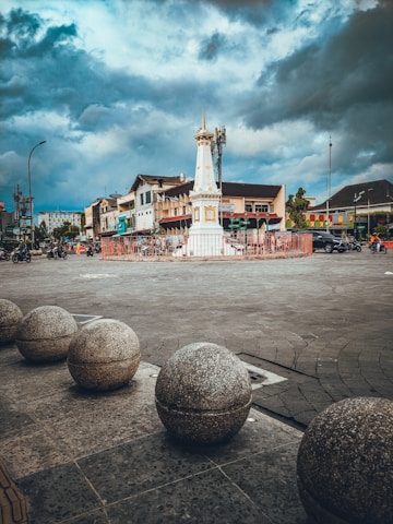 A historical monument stands in the center of a roundabout, surrounded by stone spheres that line the foreground. The sky is overcast with dramatic clouds. In the background, buildings and a few vehicles, including motorcycles, are visible. The atmosphere is a mix of urban life and historical architecture.