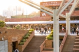 A lively university campus scene with students walking and socializing in a courtyard area. The architecture features modern building elements with brick walls and large support beams. Lush green plants are interspersed around the pathways, contributing to a welcoming atmosphere. The sunlight casts a warm glow over the scene, creating a pleasant and energetic environment.