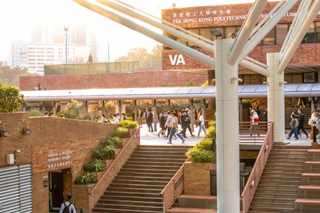 A lively university campus scene with students walking and socializing in a courtyard area. The architecture features modern building elements with brick walls and large support beams. Lush green plants are interspersed around the pathways, contributing to a welcoming atmosphere. The sunlight casts a warm glow over the scene, creating a pleasant and energetic environment.